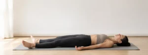 A yoga practitioner resting in Savasana (Corpse Pose) in a calm, light-filled studio. Often practiced at the end of a yoga session, Savasana encourages deep relaxation, nervous system recovery, and mindful awareness while allowing the body to integrate the effects of the practice