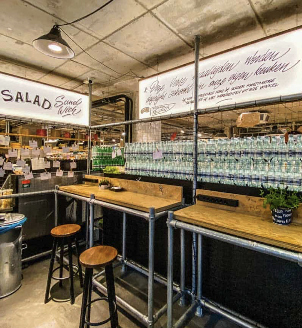 An interior view of Marqt Amsterdam showing its industrial design, seating area and neatly arranged grocery displays. The image reflects the market-style atmosphere that helped distinguish Marqt from a conventional supermarket.