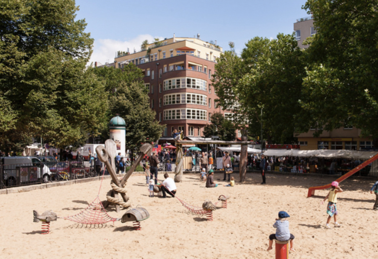 Children playing in a sandy playground in Prenzlauer Berg Berlin surrounded by trees, apartment buildings, and a neighborhood market atmosphere.