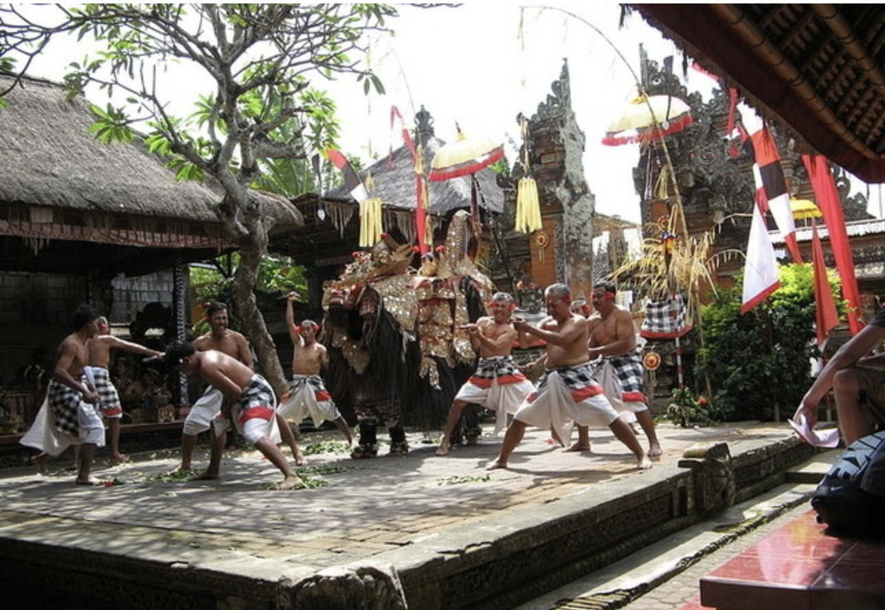 This image captures a live Barong dance performance in a Balinese temple, showcasing male dancers wearing traditional sarongs in a trance-like state, protecting their village against the evil spirit Rangda. The ornate Barong costume stands at the center, surrounded by ceremonial decorations and temple gates. A vibrant representation of Bali's cultural and spiritual heritage.