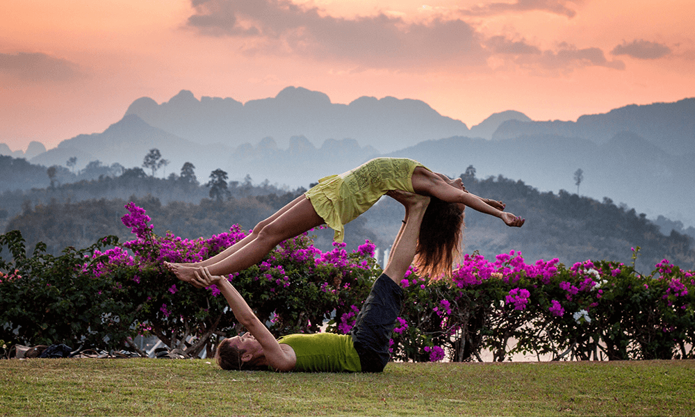 Two people in green outfits doing acroyoga in front of some flowers. bff 2 person yoga poses
