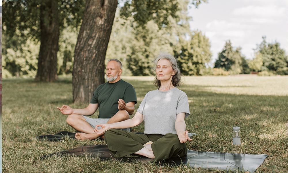 Two older people meditating in a park in front of some trees. bff 2 person yoga poses
