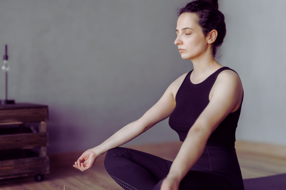 Woman sitting in a meditative yoga pose indoors with eyes closed, practicing mindfulness and calm breathing.