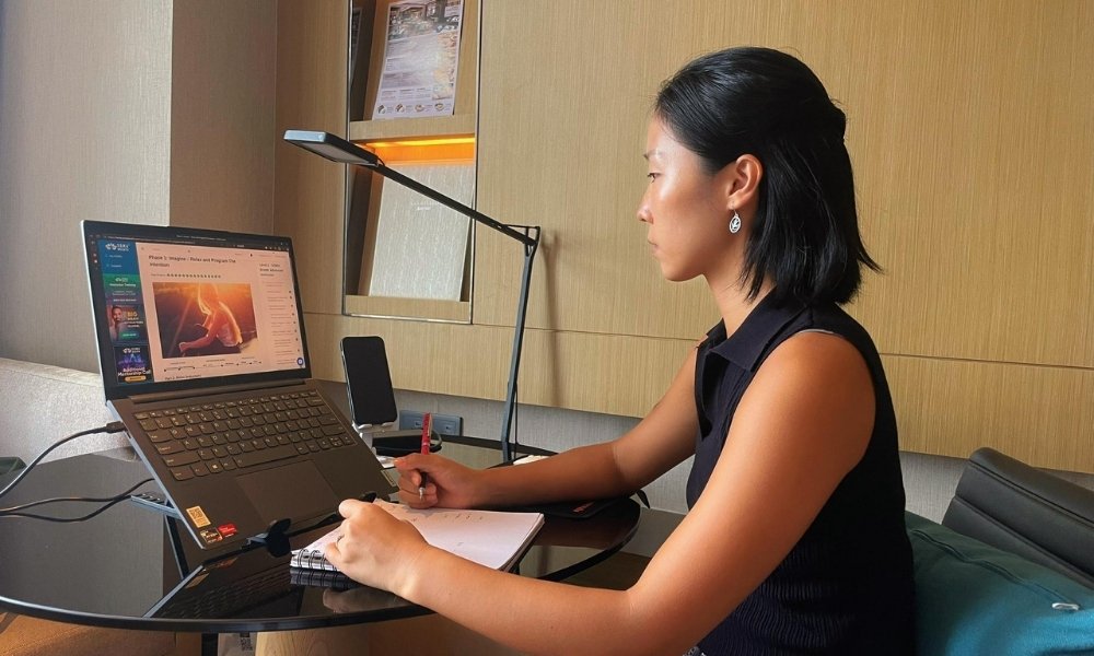Woman studying SOMA Breath Level 2 training on laptop while taking handwritten notes at a modern desk.