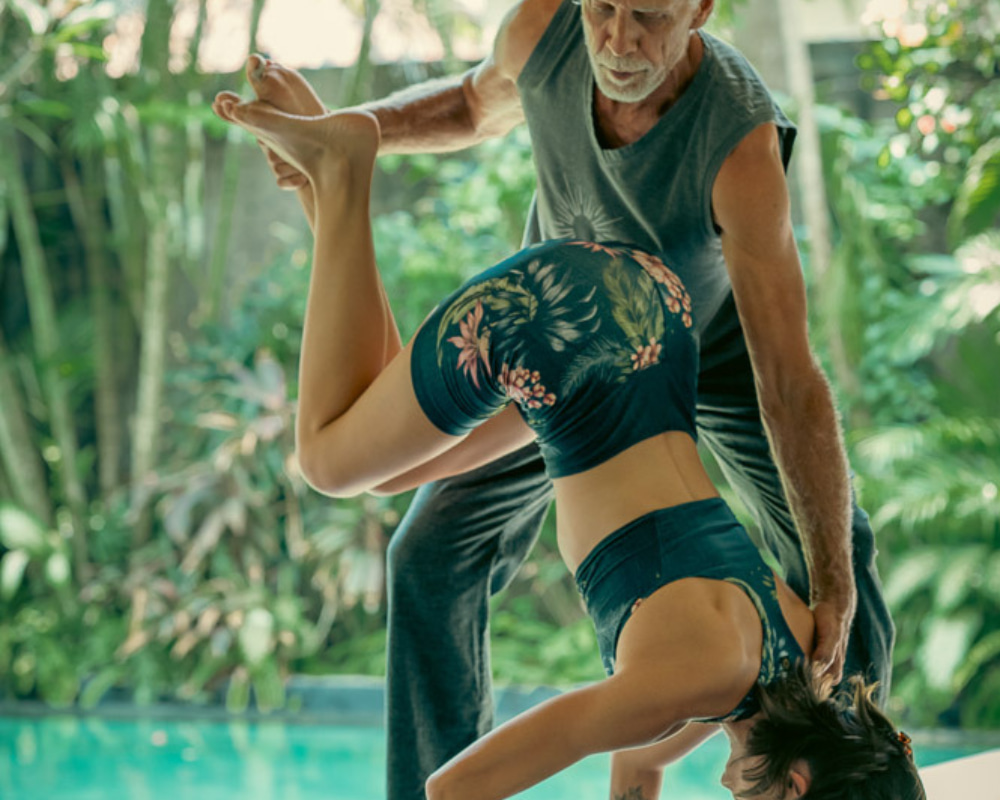 Yoga instructor assisting a student in an advanced yoga pose outdoors, surrounded by lush tropical greenery