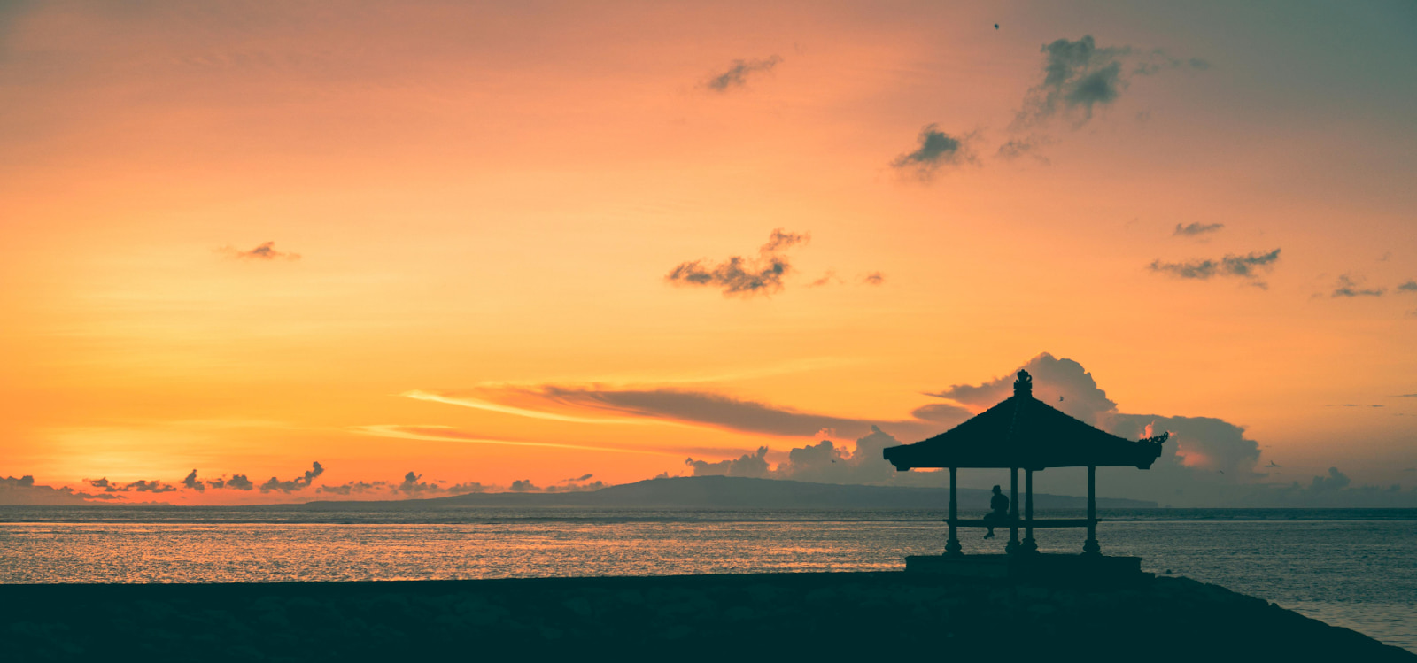 Serene sunset scene in Bali with a person enjoying the view from a seafront bale.