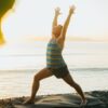 Aaron Star practicing Warrior 1 (Virabhadrasana I) yoga pose on a mat by the beach during sunrise, with vibrant ocean and sky in the background, framed by greenery