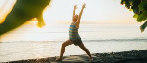 Aaron Star practicing Warrior 1 (Virabhadrasana I) yoga pose on a mat by the beach during sunrise, with vibrant ocean and sky in the background, framed by greenery