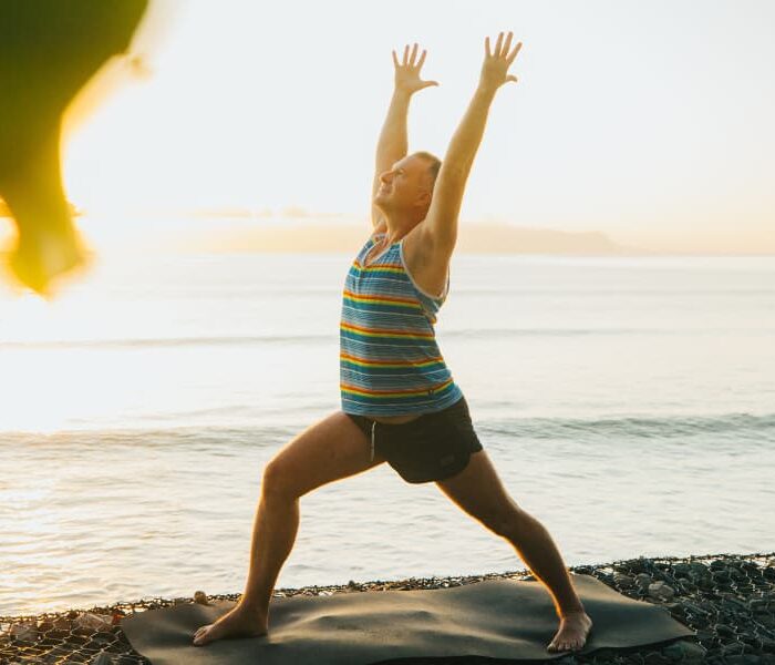 Aaron Star practicing Warrior 1 (Virabhadrasana I) yoga pose on a mat by the beach during sunrise, with vibrant ocean and sky in the background, framed by greenery