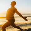 A man in Warrior 1 pose on a sunny beach at sunrise, showcasing ideal beachfront yoga locations for a Bali retreat