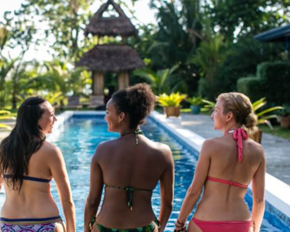 Three women in swimsuits, looking out over a pristine swimming pool at what appears to be a tropical resort, suggesting relaxation and rejuvenation at a Bali yoga retreat. Photo credit : @BlueOsaYoga