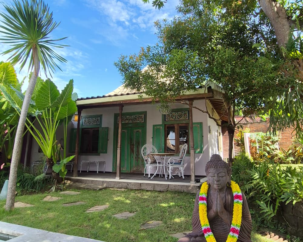Charming Balinese house with green shutters and lush garden, featuring a statue of Buddha in the front yard