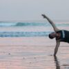 A man performs a balancing yoga pose on a beach at sunrise or sunset, symbolizing the connection with nature and the renewal of spirit found at a Bali yoga retreat.