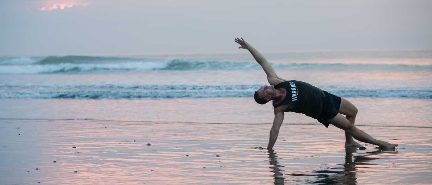 A man performs a balancing yoga pose on a beach at sunrise or sunset, symbolizing the connection with nature and the renewal of spirit found at a Bali yoga retreat.