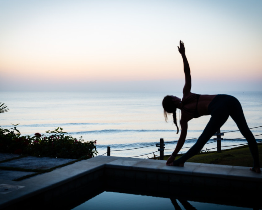 Woman practicing yoga by the ocean during sunrise, performing a triangle pose