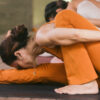 A woman in orange yoga attire performing a complex yoga pose, seated on a black yoga mat, with other blurred individuals in the background