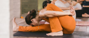 A woman in orange yoga attire performing a complex yoga pose, seated on a black yoga mat, with other blurred individuals in the background