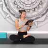 Smiling woman in lotus pose on yoga mat holding a tablet, representing online yoga practice at home.