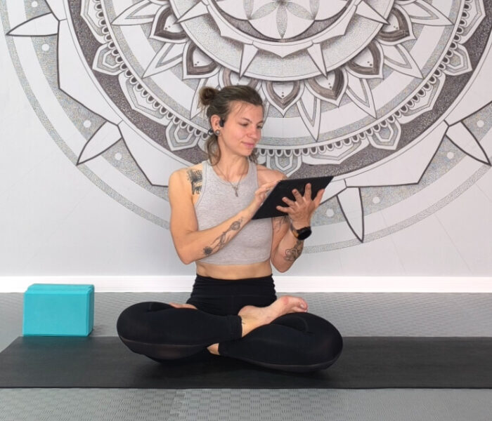 Smiling woman in lotus pose on yoga mat holding a tablet, representing online yoga practice at home.