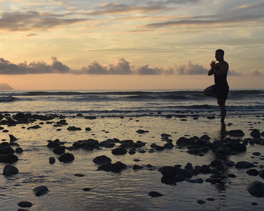 A yogi in a tree pose at sunrise on a rocky beach, showcasing beachfront yoga spaces at a Bali retreat, like Yeh Leh Bali