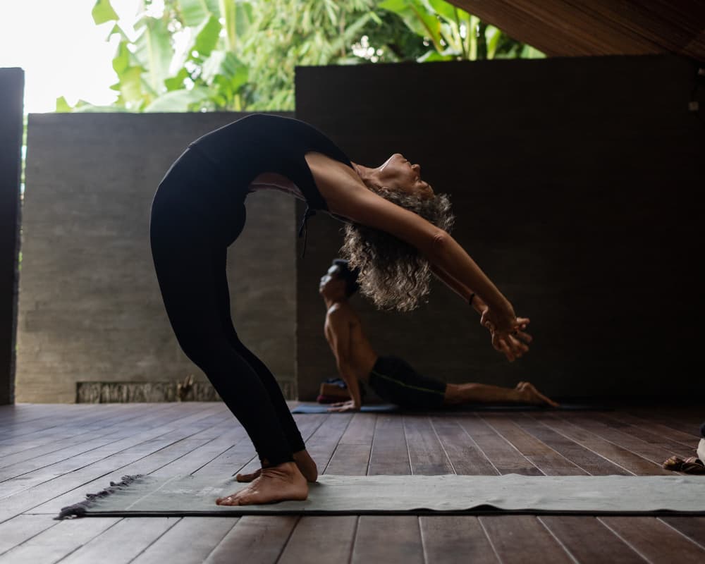 A woman in a black outfit performs a deep backbend yoga pose on a mat, with a man in a downward-facing dog pose in the blurred background. The setting appears to be an open-air yoga studio with wooden floor