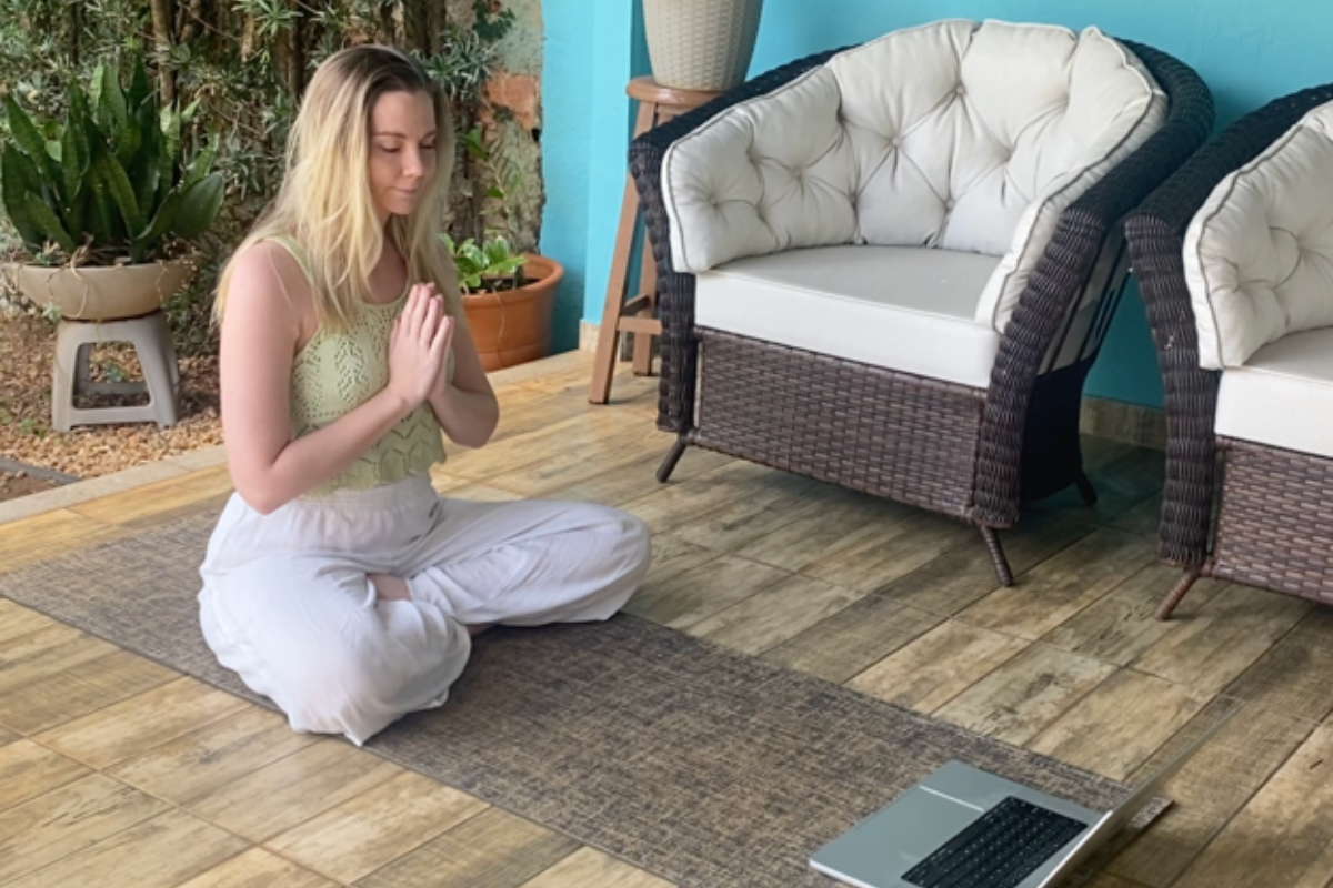 Tenille, a Sacred Path Yoga student, sits cross-legged on a yoga mat in a serene outdoor setting. With hands in Anjali Mudra (prayer position), she practices a moment of mindfulness and gratitude in front of her laptop, engaging in an online yoga teacher training session.