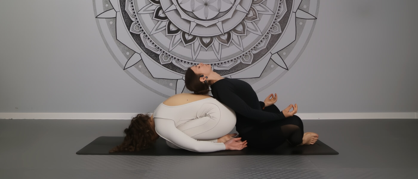 Two women practicing partner yoga for BFFs, wearing contrasting black and white outfits, with a mandala in the background.