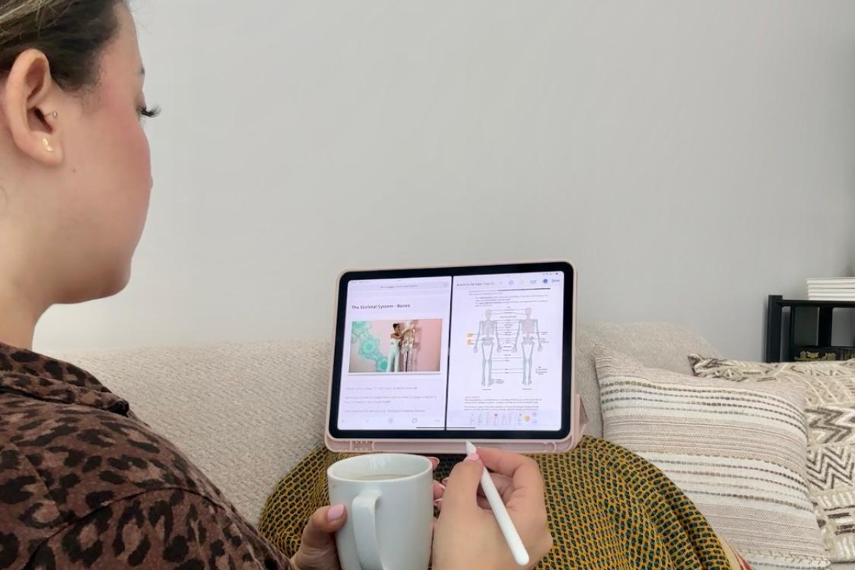 Me studying anatomy at home with her tablet, reviewing skeletal system diagrams while enjoying a warm cup of tea or coffee. Perfect setup for online yoga teacher training study sessions.