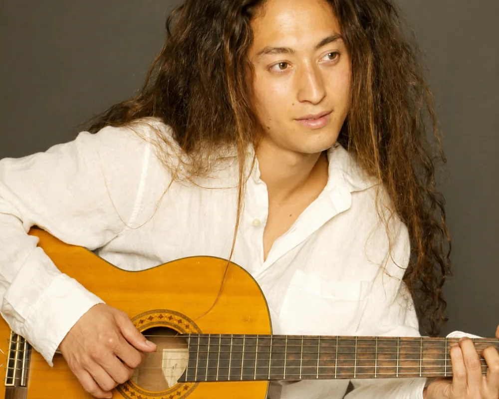 A portrait of a young Tamal Dodge playing an acoustic guitar during the period when he pursued music and visual arts before returning to yoga. Raised in an ashram in Hawaii and teaching yoga from the age of 10, Dodge explored a career in music before rediscovering his calling as a yoga teacher. Today he teaches meditation, yoga philosophy, and teacher trainings internationally after the closure of Bodhi.