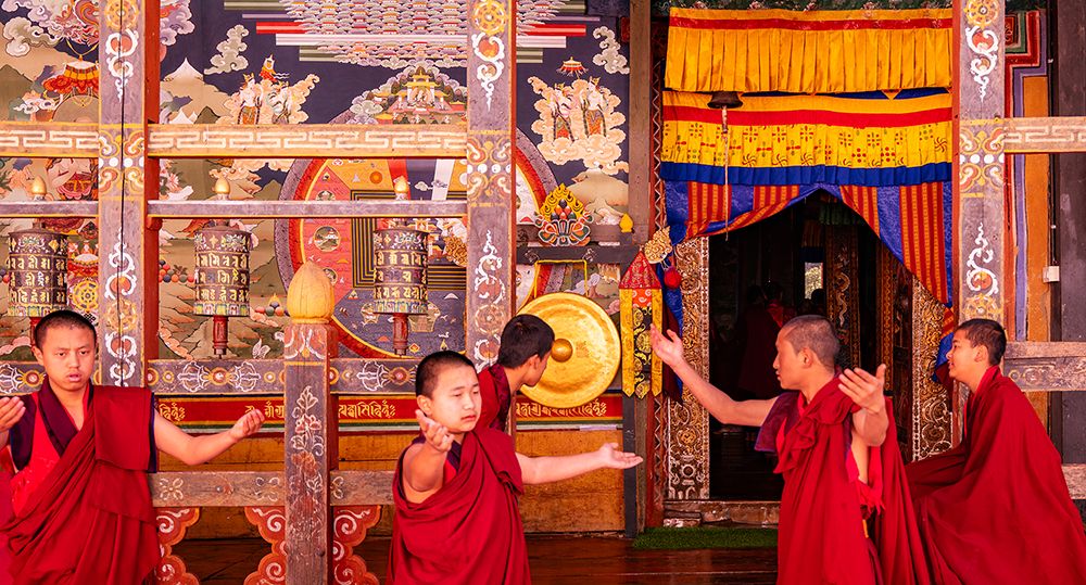 Young Buddhist monks in red robes practicing ritual gestures inside a decorated monastery in Bhutan.