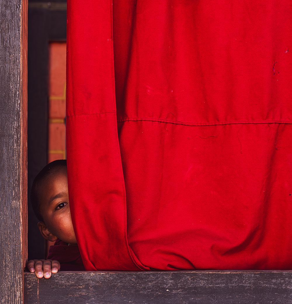Young monk peeking curiously from behind a red robe inside a Bhutanese monastery.
