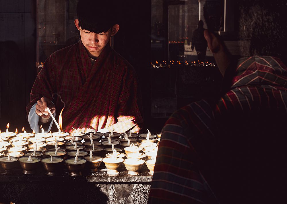 Young Bhutanese man lighting butter lamps inside a Buddhist temple in Bhutan, with warm candlelight illuminating his face and hands