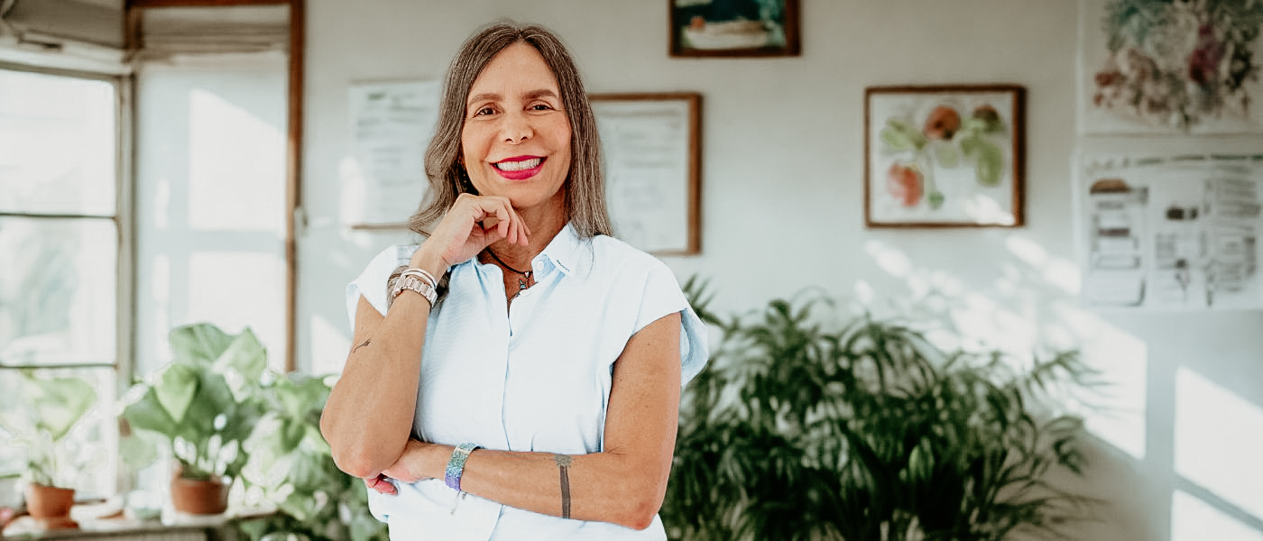 The image shows Martha Vargas, a PSYCH-K® instructor, standing confidently in a bright and natural space filled with plants. She is smiling, with a hand gently resting on her chin, and wearing a light blue button-down shirt. The background features a cozy, plant-filled room with framed artwork on the walls, exuding a welcoming and calming atmosphere. This photo captures Martha’s warmth, professionalism, and connection to both nature and the transformational work she does with PSYCH-K®. Martha Vargas PSYCH-K® instructor . PSYCH-K® transformation expert. healing and subconscious reprogramming. PSYCH-K® Bali workshop. subconscious belief change.