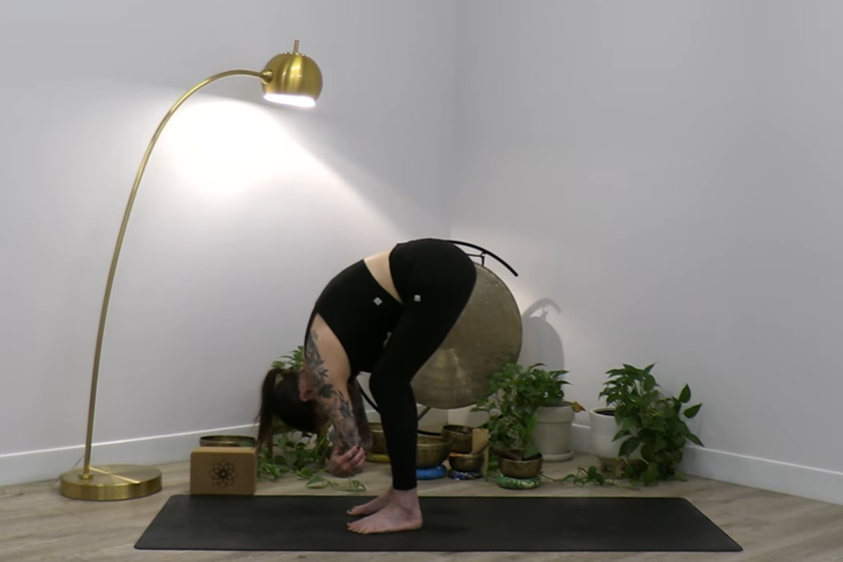 A woman in black activewear performs a standing forward fold on a yoga mat. Her upper body hangs toward the floor with arms relaxed. The space features plants, a large gong, and warm lighting, creating a peaceful yoga environment.