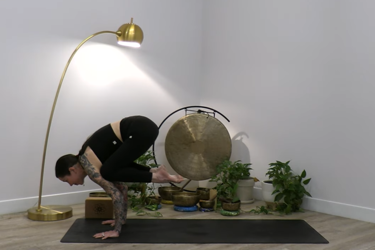 A woman demonstrates Crow Pose (Bakasana), balancing her knees on her upper arms with palms grounded on a yoga mat. The serene indoor setting includes houseplants, a decorative gong, and soft lighting.