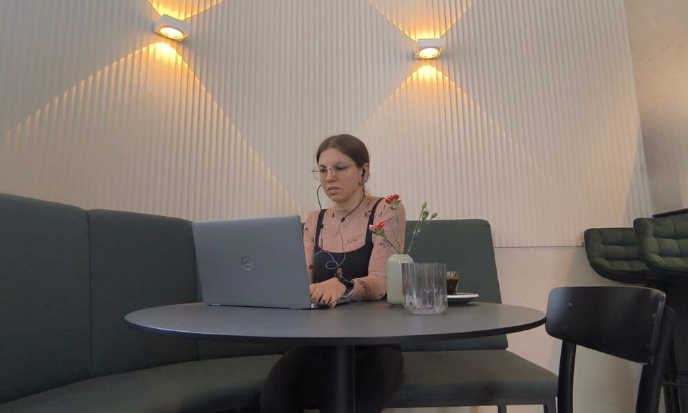 A woman sitting at a table at a cafe, working on her laptop with wired earbuds from DefenderShield/