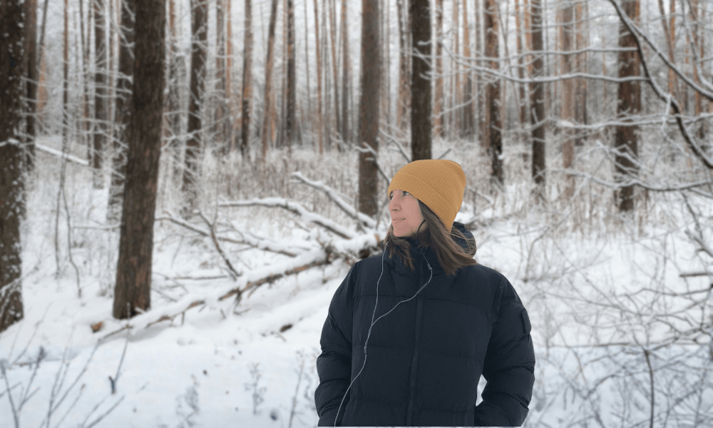 A woman wearing a black aerogel insulation jacket and a yellow hat, standing in a snowy winter forest.