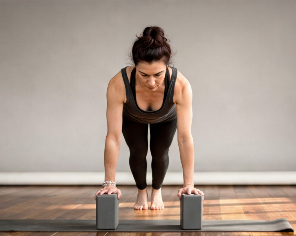 A woman practices a yoga pose using two blocks, leaning forward with both hands pressing on the blocks while standing on a mat.