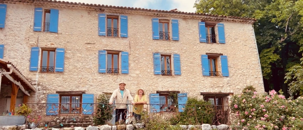 Berté et Carina posent devant la façade principale de la Ferme Saint Pierre à Bargème. La photo met en valeur l’architecture en pierre du domaine, ses volets bleus et l’atmosphère chaleureuse de cette ferme d’accueil du Haut Var.